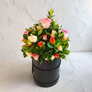 Bouquet of pastel pink and peach roses with glossy green leaves in a black round hatbox on a textured light wall backdrop