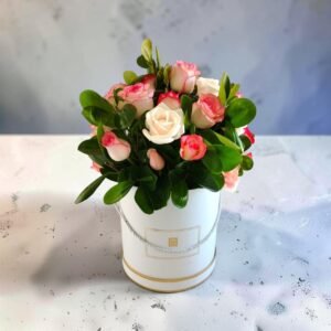 Bouquet of pink and white roses with glossy green leaves arranged in a white cylindrical gift hat box with a silver rope handle and gold trim.